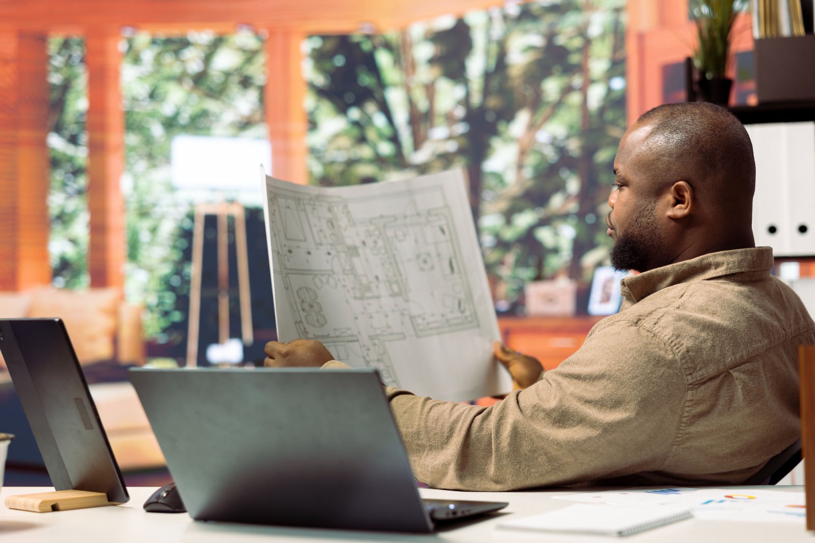 Texto alternativo sugerido: Homem sentado à mesa em um escritório, analisando uma planta arquitetônica impressa. À sua frente há um notebook aberto, papéis espalhados e um ambiente bem iluminado com janelas grandes e vegetação visível ao fundo.